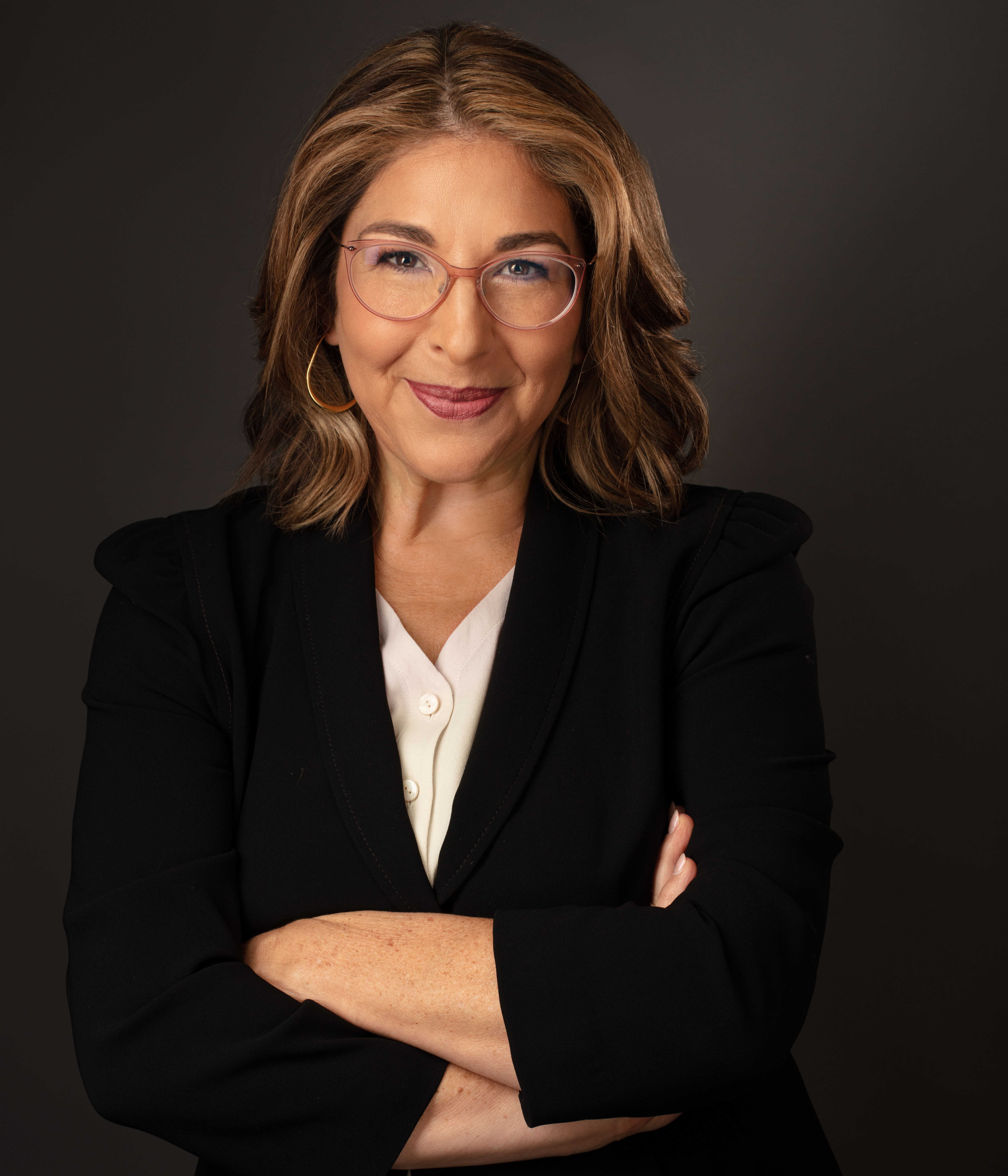 Naomi is a white woman with shoulder length brown hair and blonde hair. She is smiling at the camera with her arms folded. She wears glasses, a white shirt and a dark blazer.