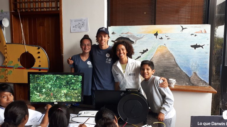 Salome stands in a classroom with her arms around two other members of her team, and a schoolchild. Behind them is a poster of a seamount. Other children are pointing at an underwater image on a screen,