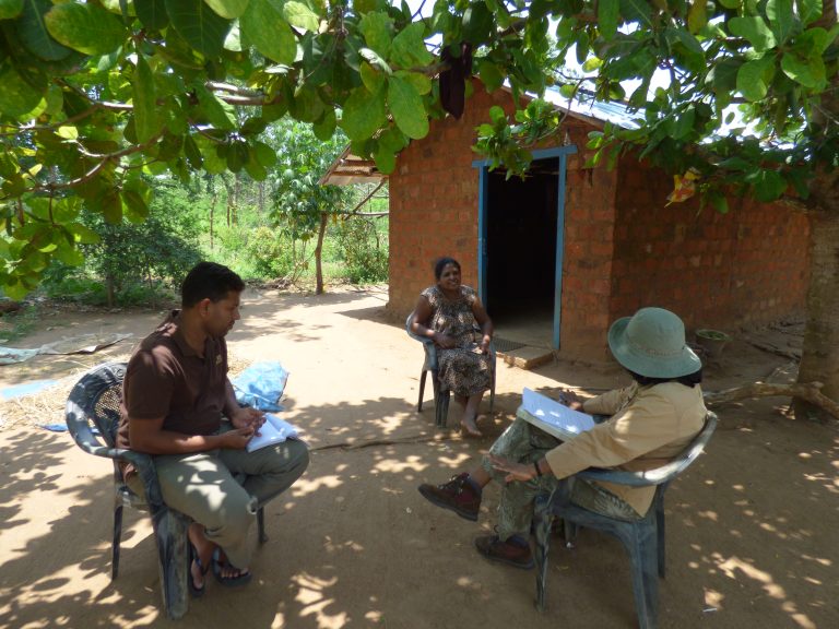 A researcher sits outdoors speaking with two other people, sitting on chairs under a tree.