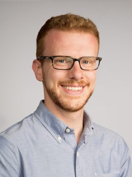 A young man with red hair, beard and moustache, wearing dark rimmed glasses.