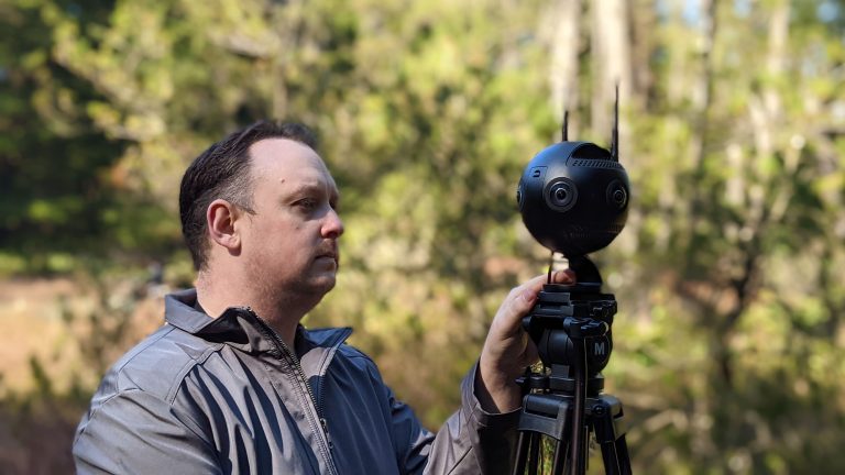A white man stands with his hand on a 360 camera - which looks like a black ball with antennae mounted on a tripod. Bog foliage is out of focus in the background.