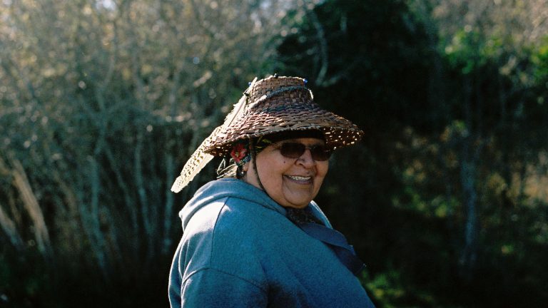 A woman in a traditional woven hat, sunglasses and blue hoodie smiles at the camera