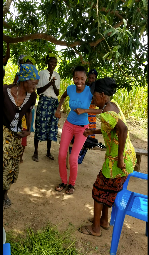 Jemima dances with a group of Ghanaian women after a research sessoin. They are outdoors, under some trees.