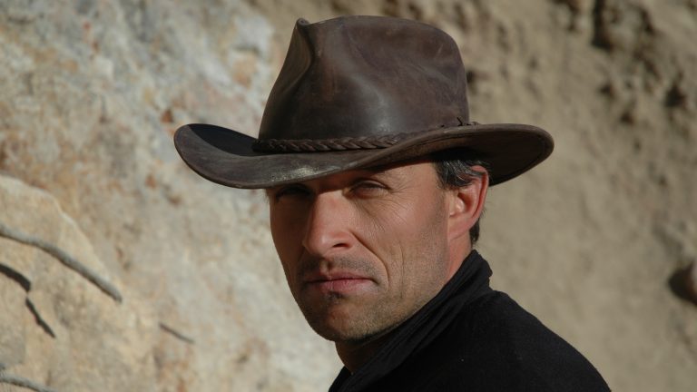 Matthias is looking towards the camera (and the sun!) in a black turtle neck shirt and a brown hat with a wide brim.