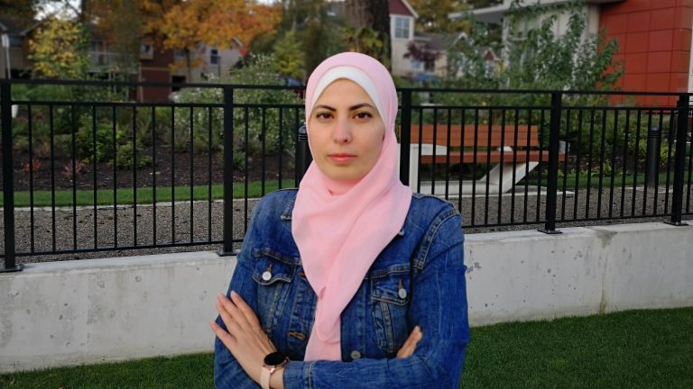 Nisreen stands in front of a suburban setting with fall foliage. She is wearing a denim jacket and pink head scarf, standing with her arms folded.