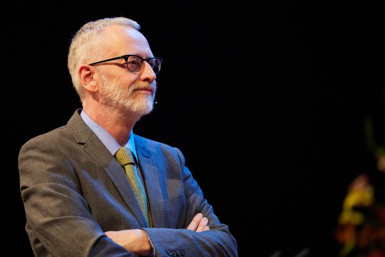 Dan is a white man with short white hair and a closely cropped beard. He wears glasses with dark frames and is standing in a suit with his arms folded.