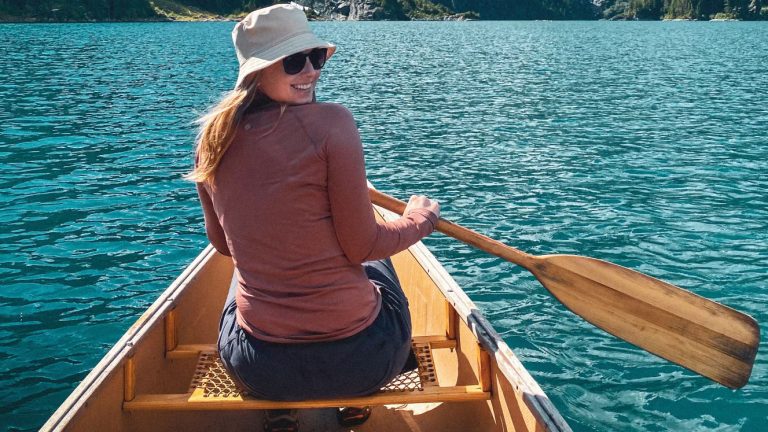 Courtney is a young white woman with long blonde hair. She is in a boat on a clear, blue mountain lake, looking back over her shoulder at the camera and holding a wooden oar. She is wearing a bucket hat and sunglasses, and a long sleeved shirt.