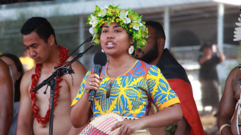 A Pacific Islander wears a lei / hei / flower crown and a patterned blue and yellow shirt. They are speaking into a microphone.