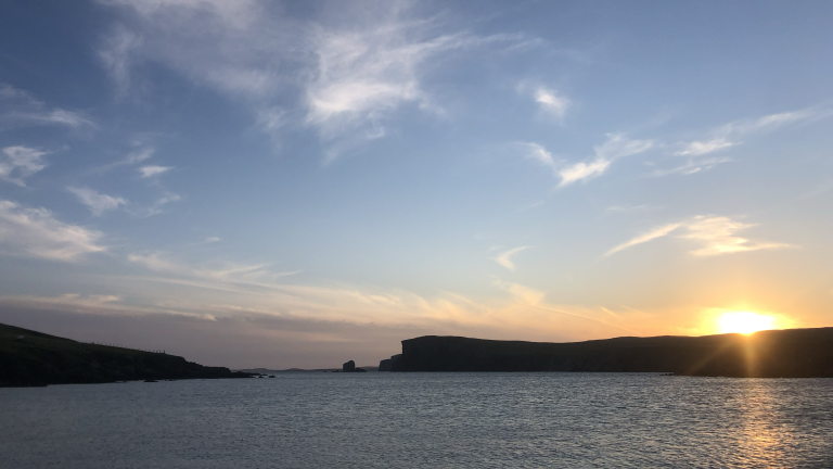 The sun sets behind a headland in the distance, with the ocean in the foreground