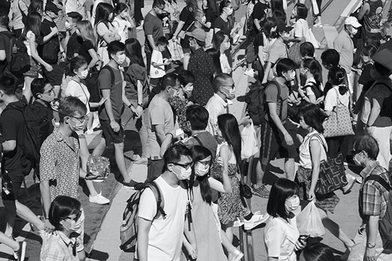 Monochrome image of a busy street with people walking in all directions, masked