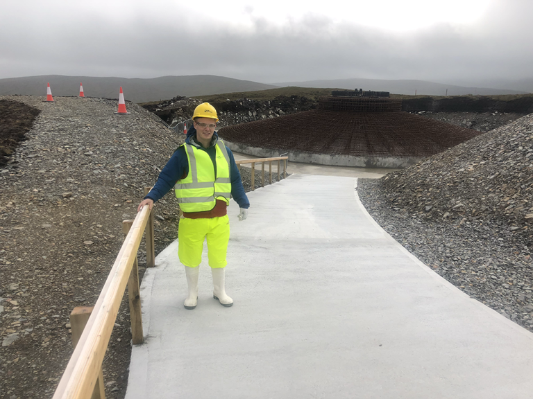 Max is a white man wearing fluorescent PPE. He stands on a concrete walkway, with the large concrete base of a wind turbine partially constructed behind him