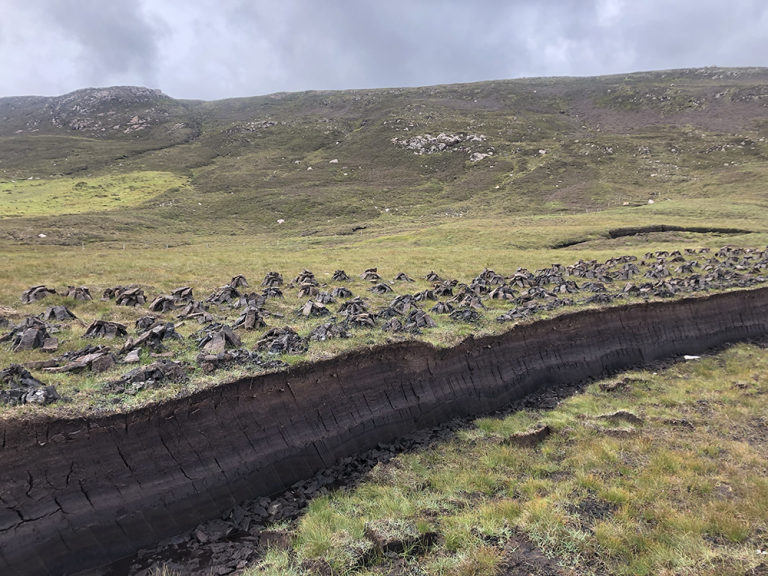 A dark strip of peat cuts horizontally across the image. Behind it, harvested segments are stacked to dry. Low hills and a grey sky lie in the background.